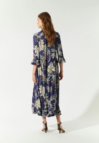 Luftiges, navyblaues Kleid mit floralem Druck, mit Rüschensäumen und -saum. Der Stoff hat eine glatte Textur und reicht bis zur Mitte der Waden.