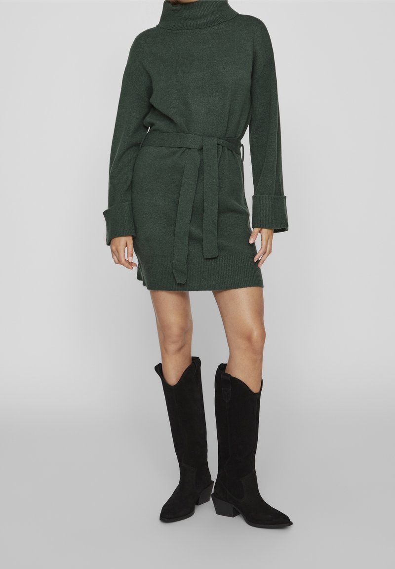 Femme portant une robe en maille ceinturée vert foncé et des bottes à talons noires mi-mollet, debout sur un fond gris.