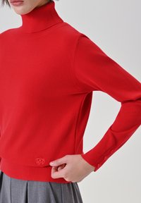 Roter Strickpullover mit Rollkragen, langen Ärmeln, geripptem Kragen und seitlichen Schlitzen. Mit dekorativen Knöpfen an den Ärmeln, weicher Textur.