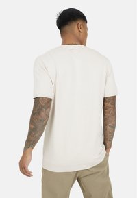 Kremowa, dzianinowa koszulka z krótkim rękawem, okrągłym dekoltem; posiada prążkowane akcenty i luźny krój, pokazana z tyłu z widocznymi tatuażami.