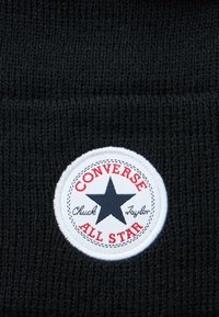 Zwart gebreid stof met een witte ronde patch waarop een zwarte ster en rode tekst "Converse All Star" staat met "Chuck Taylor" in cursief.