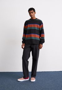 Black long-sleeve shirt with orange, green, and blue horizontal stripes, paired with loose dark jeans and red-and-white checkered slip-on shoes.