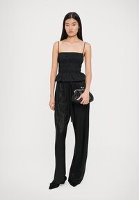 Top negru peplum cu bretele subțiri, asortat cu pantaloni largi texturați negri. Modelul ține în mână o mică poșetă neagră.