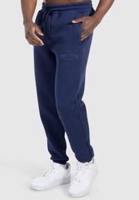 Marinefarbene Sweatpants mit elastischem Bund und Kordelzug, ausgestattet mit Seitentaschen und gesticktem Logo auf dem linken Oberschenkel. Weiße Sneakers.