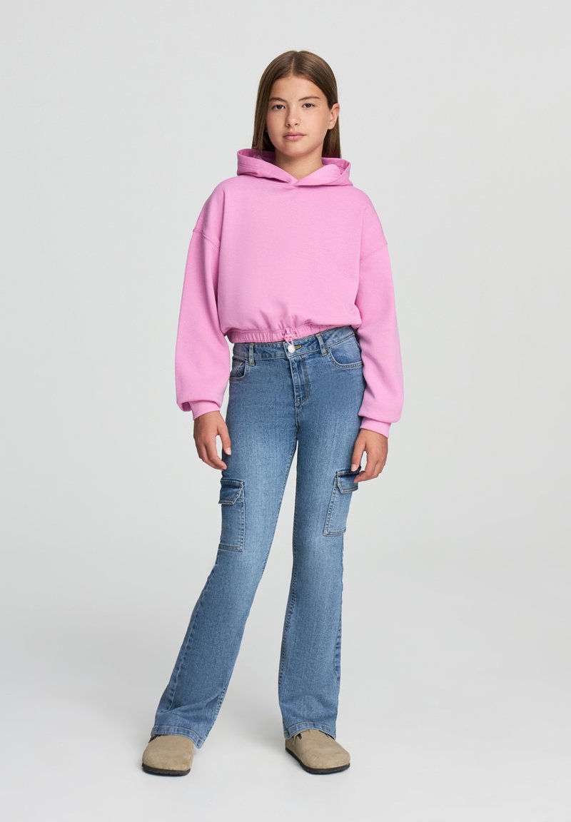 Felpa rosa con coulisse e orlo elastico abbinata a jeans cargo a zampa d'elefante di un blu chiaro con tasche laterali. Le calzature sono slip-on beige.