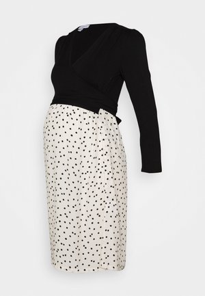 Haut cache-cœur noir à manches longues avec une jupe blanche à pois noirs irréguliers dans une robe de maternité longueur genou.