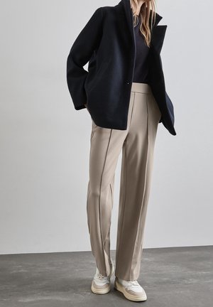 Cappotto in misto lana blu navy con un design strutturato, abbinato a pantaloni beige su misura e sneakers bianche con dettagli sottili.