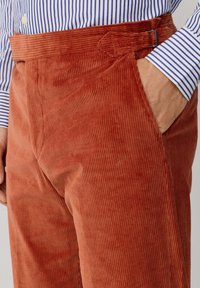 Corduroy pants in rust orange with vertical ribbing, featuring a side pocket and a belt loop, paired with a blue and white striped shirt.