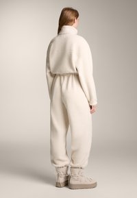 Kremno obarvan flis kombinezon z visokim ovratnikom, elastičnim pasom in zoženimi hlačnicami. Ima mehak teksturo in rebraste manšete.
