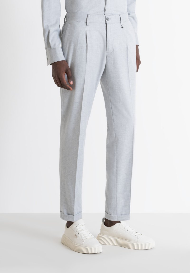 Pantaloni grigi chiari su misura con pieghe, fondo con risvolto, tasche laterali, abbinati a sneaker bianche, caratterizzati da un tessuto testurizzato e cuciture ordinate.