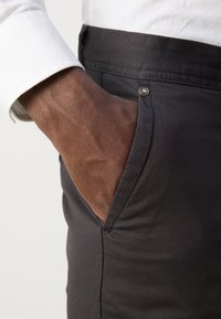 Pantalons noirs en tissu tissé, avec une poche latérale et un accent en bouton en bronze à la taille. Gros plan d'une main dans la poche.