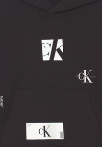 Felpa nera con cappuccio caratterizzata da un logo bianco prominente con la scritta "CK" e "Calvin Klein Jeans", tasca frontale e tessuto dalla texture liscia.