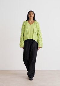 Πλεχτό καμπαρντίνα σε φωτεινό λάιμ πράσινο με σχέδιο σε καλώδιο, φορεμένη πάνω από ένα ταιριαστό τοπ με V. Συνδυασμένο με φαρδιά μαύρα παντελόνια και παπούτσια.