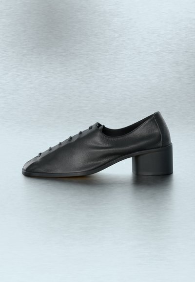 Μαύρο δερμάτινο παπούτσι με χαμηλό τακούνι και στρογγυλεμένη μύτη, διακοσμητικά κουμπιά στο επάνω μέρος και τακούνι σε σχήμα τετραγώνου, τοποθετημένο πάνω σε βουρτσισμένη μεταλλική επιφάνεια.