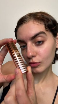 Zylindrische Lippenstiftkapsel in Roségold mit vertikalen Rillen, zwischen den Fingern gehalten. Hintergrund ist einfarbig und hell.