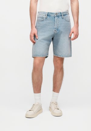 Mężczyzna ubrany w jasnoniebieskie jeansowe szorty, białe skarpetki i off-white sneakersy, stojący na jednolitym tle.