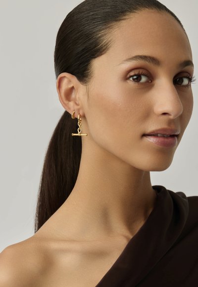 Pendientes de oro con un diseño de barra y detalle de eslabones de cadena, colgando de un aro circular. La modelo tiene el cabello largo y liso.