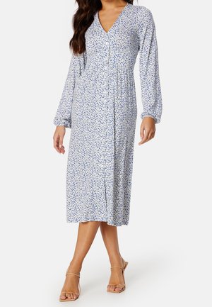 Kobieta w sukience midi z długim rękawem, dekoltem w serek i zapinanej na guziki, z niebiesko-białym kwiatowym wzorem, w połączeniu z beżowymi sandałami na obcasie z odkrytymi palcami.