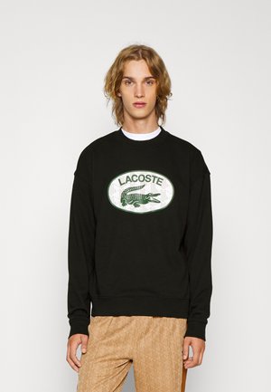 Sweatshirt noir avec un col rond, arborant un grand patch logo Lacoste vert et blanc au centre. Texture de tissu douce.
