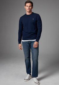 Maglione blu navy a maglia con scollatura rotonda, indossato sopra una camicia bianca. Abbinato a jeans blu a gamba affusolata e sneakers bianche con dettagli grigi.