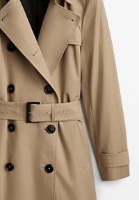 Trench-coat beige avec un design croisé, de grands boutons noirs, une ceinture avec une boucle carrée et un col cranté. Tissu texturé.