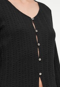 Μαύρη υφασμάτινη ζακέτα με βαθύ ντεκολτέ σε σχήμα V, με λαμπερά ασημένια κουμπιά μπροστά και περίπλοκο υφαντό μοτίβο σε όλο το ύφασμα.