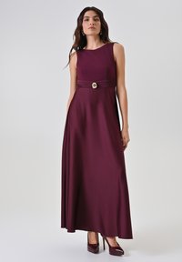 Robe maxi sans manches bordeaux avec un corsage ajusté, une taille plissée et un bouton décoratif doré. Tissu lisse avec un léger éclat.