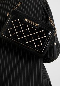 Czarna torebka na ramię z teksturowanym panelem, złotymi nitami i sztucznymi perłami. Łańcuszkowy pasek i dekoracyjny frędzel po boku.