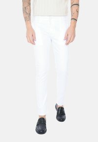 Pantaloni bianchi slim-fit con una texture liscia, dotati di chiusura con bottone e zip, abbinati a mocassini neri.