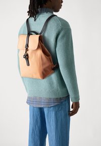 Brun lærersekk med en fold-over klaff og svarte aksentstropper, båret over en teksturert lyseblå genser og løse denimbukser.