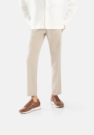 Sneaker in pelle marrone con suole bianche, pantaloni beige chiaro su misura e una camicia bianca a bottone, con una tasca a sinistra.