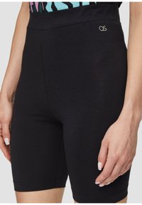 Gros plan sur une personne portant un short noir moulant mi-cuisse avec un petit logo "QS" sur la taille, la main reposant sur le côté.