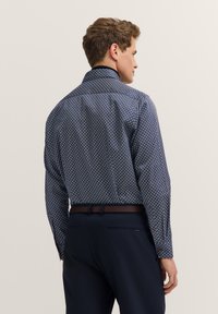 Camicia blu navy con piccolo motivo geometrico bianco, colletto con bottoni, maniche lunghe e una finitura di tessuto liscia, indossata con pantaloni scuri.