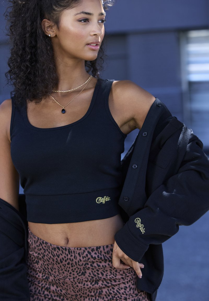 Black ribbed crop top with yellow logo, paired with a black oversized jacket, worn over leopard-patterned leggings. Smooth texture throughout.