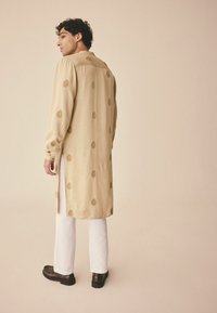 Beige Langarmhemd mit goldenen, kreisförmigen Mustern, Knopfmanschetten und seitlichen Schlitzen, kombiniert mit weißen Hosen und dunkelbraunen Schuhen.