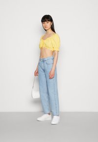 Top amarelo cropped com mangas bufantes, combinado com jeans de cintura alta azul claro. Ténis brancos e uma pequena mala branca complementam o visual.