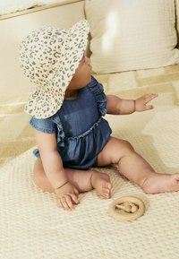 Robe en denim avec des manches volantes, chapeau seau beige avec un imprimé léopard vert, et un jouet en bois en forme d'anneau sur un tapis clair texturé.