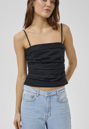 Frau trägt ein schwarzes, gerafftes Camisole mit dünnen Trägern und hellblaue, hoch taillierte Jeans vor einem schlichten grauen Hintergrund.