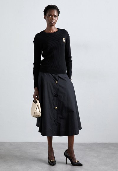 Pull noir côtelé avec un petit logo, associé à une longue jupe noire à boutons dorés. Des escarpins noirs et un sac à main crème complètent le look.