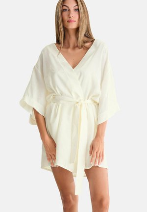 Femme aux longs cheveux blonds portant une robe ample de couleur crème ceinturée, aux manches larges, se tenant devant un fond clair uni.