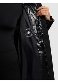 Melna virsjaka ar spīdīgu oderi, logotipu, rāvējslēdzenes aizdari un iekšējām kabatām, kas izceļ gludu tekstūru un modernu dizainu.