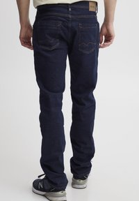 Vaqueros de mezclilla azul oscuro con un corte recto, que presentan dos bolsillos traseros con solapas puntiagudas y costuras en contraste. Llevados con zapatillas deportivas.