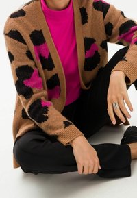 Brun kofta med svarta och rosa hjärtan, med en avslappnad passform och långa ärmar, bärs över en ljusrosa topp och svarta byxor.