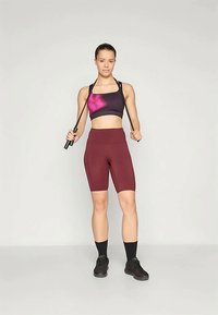 Svart sports-bh med rosa tie-dye-design, kombinert med høyt skårne burgunder shorts. Modellen holder et hoppetau og har på seg svarte sneakers og sokker.