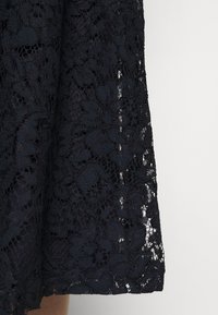 Černá krajková látka s květinovým vzorem a průsvitnými květinovými vzory na pevném podkladu, zobrazující okraj lemu na světlém pozadí.