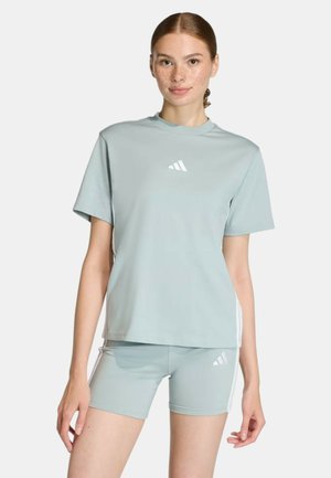 Joven mujer vistiendo una camiseta azul claro de Adidas y pantalones cortos a juego con el logo, posando contra un fondo blanco liso.