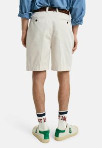 Pantalones cortos color crema con un corte recto, que cuentan con dos bolsillos traseros y un cinturón trenzado marrón. Combinados con zapatillas blancas y verdes y calcetines con estampado.