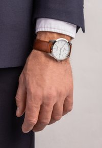 Montre avec bracelet en cuir marron, boîtier en acier inoxydable argenté, cadran blanc, index horaires noirs et guichet de date à 3 heures.