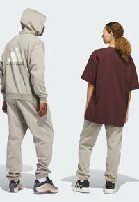 Helles graue Hoodie mit großem weißen Logo, kombiniert mit passenden Jogginghosen. Burgunderfarbener Oversized-T-Shirt und beigefarbene Jogginghosen. Beide tragen sportliche Schuhe.