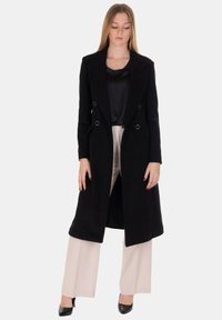 Cappotto lungo nero con design doppiopetto, texture liscia, grandi rever e dettagli in metallo color argento, abbinato a pantaloni crema a gamba larga.